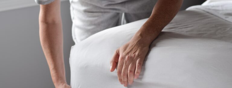 Eine Person bezieht das Bett mit einem frischen weißen Spannbettlaken – Symbol für erholsamen Schlaf trotz nächtlichem Schwitzen.