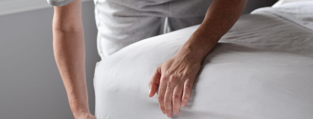 Eine Person bezieht das Bett mit einem frischen weißen Spannbettlaken – Symbol für erholsamen Schlaf trotz nächtlichem Schwitzen.